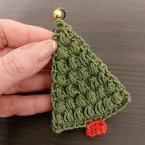 May include: A green crocheted Christmas tree ornament with a red crocheted star at the bottom. The ornament has a gold bead at the top.