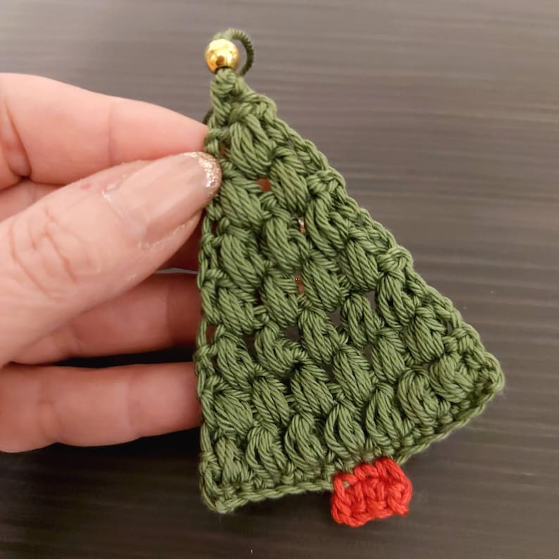 May include: A green crocheted Christmas tree ornament with a red crocheted star at the bottom. The ornament has a gold bead at the top.