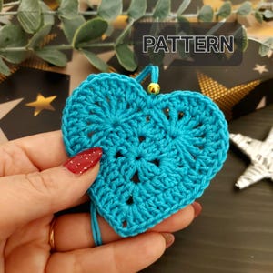 Puede incluir: Un adorno de corazón tejido a crochet en un color azul turquesa brillante. El adorno tiene una cuenta dorada en la parte superior y se sostiene en una mano.