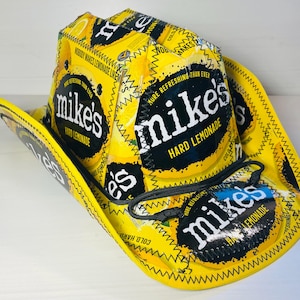 Puede incluir: Un sombrero de vaquero amarillo hecho con envases de Mike's Hard Lemonade. El sombrero presenta el logotipo y el texto de la marca, con costuras negras y una correa de barbilla negra. El ala y la corona son amarillas.