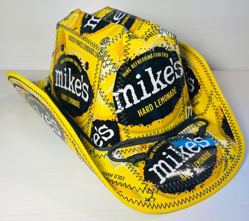Puede incluir: Un sombrero de vaquero amarillo hecho con envases de Mike's Hard Lemonade. El sombrero presenta el logotipo y el texto de la marca, con costuras negras y una correa de barbilla negra. El ala y la corona son amarillas.