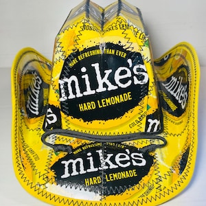 Puede incluir: Un sombrero de vaquero amarillo hecho con envases de Mike's Hard Lemonade. El sombrero presenta el logotipo de Mike's Hard Lemonade en blanco y negro, con las palabras "HARD LEMONADE" debajo. El sombrero tiene una correa de barbilla negra y est&aacute; cosido con hilo amarillo.