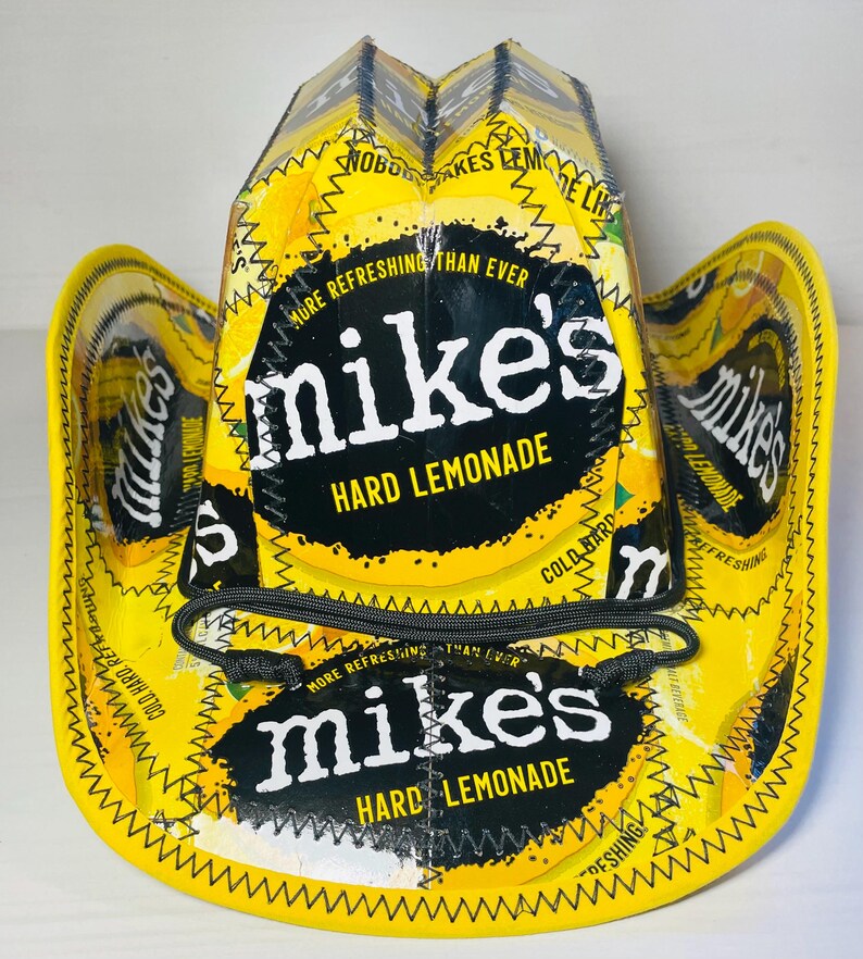 Puede incluir: Un sombrero de vaquero amarillo hecho con envases de Mike's Hard Lemonade. El sombrero presenta el logotipo de Mike's Hard Lemonade en blanco y negro, con las palabras "HARD LEMONADE" debajo. El sombrero tiene una correa de barbilla negra y est&aacute; cosido con hilo amarillo.