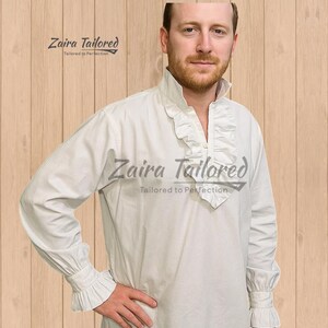 Op de afbeelding: Een wit overhemd met lange mouwen en een kraag en manchetten met ruches. Het overhemd heeft een knoopsluiting aan de voorkant en is gemaakt van een zachte, soepele stof. De tekst "Zaira Tailored" is op het overhemd gedrukt. Het is een historisch of theatraal kledingstuk.