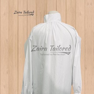 Op de afbeelding: Wit overhemd met lange mouwen, hoge kraag en manchetten met ruches. De tekst "Zaira Tailored" en "Tailored to Perfection" staat in grijs. Het overhemd is van lichte stof, op een houten achtergrond.