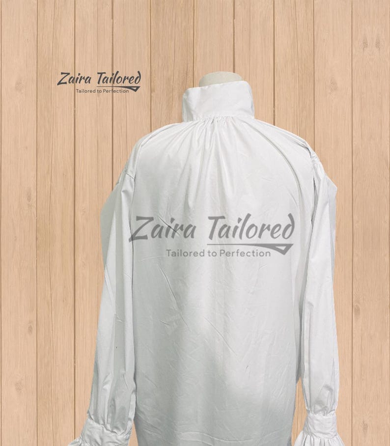 Op de afbeelding: Wit overhemd met lange mouwen, hoge kraag en manchetten met ruches. De tekst "Zaira Tailored" en "Tailored to Perfection" staat in grijs. Het overhemd is van lichte stof, op een houten achtergrond.