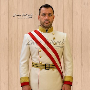 May include: A cream-colored military-style jacket with gold buttons, trim, and a black belt. A red and white sash is draped across the front. The text "Zaira Tailored" is visible.