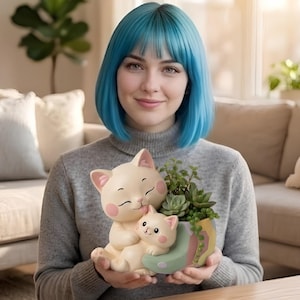 May include: A beige cat-shaped planter holding succulents. The planter features a mother cat embracing a kitten. The planter has a light green and yellow rim. The person holding the planter is wearing a gray sweater.