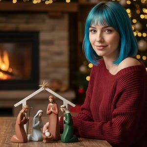 May include: A Christmas nativity scene with figurines of Mary, Joseph, and baby Jesus under a white wooden shelter with a gold star. The figurines are on a wooden table. A person with blue hair and a red sweater is sitting next to the nativity scene.