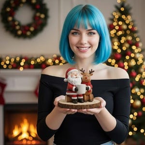 May include: A woman with blue hair holds a Christmas ornament depicting Santa Claus and a reindeer. The figures are on a wooden base. The woman wears a black off-the-shoulder top and blue jeans. A Christmas tree and fireplace are in the background.
