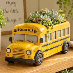 Pode incluir: Um vaso de flores amarelo em forma de ônibus escolar cheio de suculentas. O vaso tem rodas pretas, uma grade dianteira e as palavras "SCHOOL BUS" na frente. O vaso está em uma superfície de madeira.