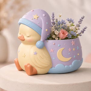 May include: A planter shaped like a sleeping yellow duck wearing a lavender nightcap with a blue brim. The planter features a lavender base decorated with a yellow crescent moon, pink and blue stars, and light blue clouds, filled with small flowers.