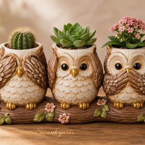 May include: A decorative planter featuring three cream and brown owl figures perched on a brown log base with small pink and blue flowers. The owls are arranged in a see-no-evil, hear-no-evil, and speak-no-evil pose, each holding a small plant.