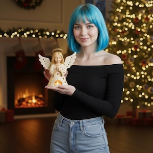 May include: A cream-colored angel figurine with outstretched wings, holding a candle holder. The angel has a halo and is wearing a robe. The candle holder has heart-shaped cutouts. The background includes a Christmas tree and a fireplace.