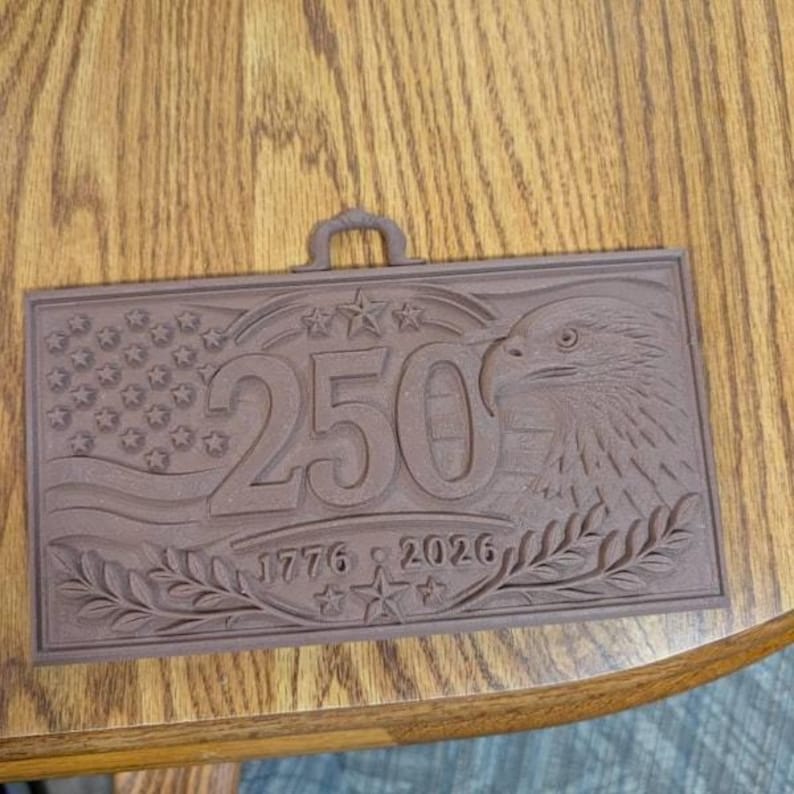 May include: A rectangular brown plaque featuring a raised design of a bald eagle, the number 250, and the years 1776 and 2026. The background includes a stylized American flag with stars and stripes, and a small hanging loop at the top center.