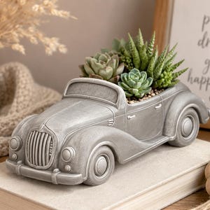 May include: A grey, vintage car-shaped planter filled with various green succulents. The planter is made of a cast material and sits on a neutral-coloured book. The car has detailed features like a grill, headlights, and wheels. The background includes a framed sign with text.