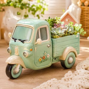 May include: A mint green ceramic planter shaped like a vintage three-wheeled truck. The truck bed is filled with succulents. The vehicle has cream-colored accents, a floral decal, and black tires. The planter is a decorative item for home or garden.