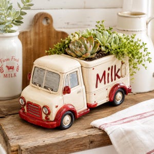 Könnte beinhalten: Cremefarbener Milchwagen-Blumentopf aus Keramik mit roten Akzenten. Der LKW-Aufbau ist mit Sukkulenten und Grünzeug gefüllt. Das Wort "Milk" ist rot geschrieben. Dekoration für das Zuhause.