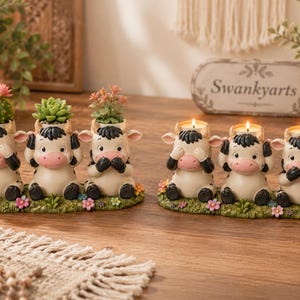 May include: Two sets of three cream-coloured cow figurines on a green grassy base with flowers. The left set holds a cactus and two succulents in the cow heads. The right set features three lit tea light candles placed inside the cow heads.