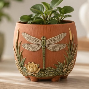 May include: A terracotta-coloured ceramic planter featuring a raised relief design of a green dragonfly, a yellow water lily, and bulrushes. The round pot has a textured surface and contains a small green plant with rounded leaves.