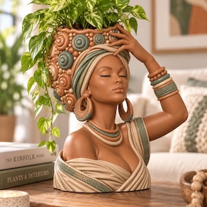 May include: A decorative bust planter featuring a person with closed eyes, wearing a headwrap adorned with circular patterns in shades of brown and teal. The figure wears hoop earrings, layered necklaces, and stacked bracelets, with a trailing green plant.