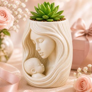 May include: A cream-colored ceramic planter featuring a mother and child design. The planter holds a green succulent plant. The mother's face is sculpted with closed eyes, cradling a sleeping baby. The background includes pink roses and a gift box.