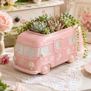 May include: A pink ceramic campervan planter filled with succulents. The planter has a retro design with flower details and white window frames. The campervan is on a white lace tablecloth.