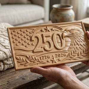 A rectangular wooden plaque with carved details. The design includes the number "250", an American flag, an eagle, and the dates "1776" and "2026". The plaque is held in front of a neutral background.