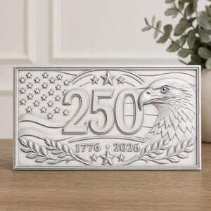 May include: A rectangular silver-toned plaque featuring a relief design of an American flag, a bald eagle head, and the number 250. The text 1776 and 2026 is centered below the number, flanked by stars and laurel leaf branches at the bottom edge.