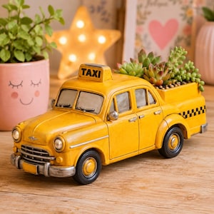 May include: A yellow taxi cab planter with a black and white checkered pattern on the back. The planter is filled with green succulents. The word "TAXI" is displayed on the roof of the cab.