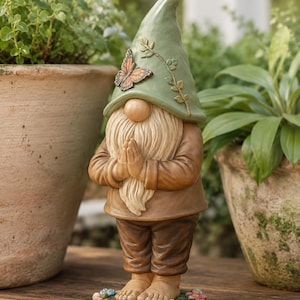 Könnte beinhalten: Eine Gartenzwerg-Figur mit einer salbeigrünen Zipfelmütze, die mit einem Schmetterling und Ranken verziert ist. Der Zwerg hat einen langen cremefarbenen Bart, trägt eine braune Tunika und Hose und steht barfuß auf einem grünen Sockel mit Blumen.
