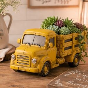 May include: A yellow vintage truck planter filled with various succulents. The truck has a distressed finish, with a slatted bed. The truck is sitting on a wooden surface. A ceramic pitcher with flowers is in the background.