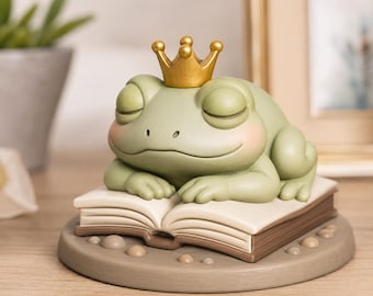 May include: A light green frog figurine with closed eyes and pink cheeks, wearing a small gold crown. The frog rests its arms on an open book, which is set upon a circular base decorated with small, smooth, round stones in a neutral colour.