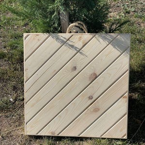 May include: A square wooden board with a diagonal pattern of wood planks. The board is light brown in color and has a smooth surface.