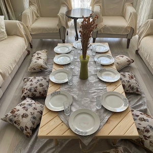 以下が含まれることがあります： 食事のためにセットされた低い木製のテーブル。花柄のクッションに囲まれています。白いお皿とグラスが灰色のランナーの上に配置されています。2つのアームチェアはベージュの布で覆われています。