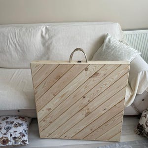 May include: A wooden folding table with a diagonal wood grain pattern. The table has a rope handle and is closed.