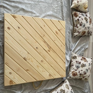 May include: A light-colored wooden tray with diagonal slats. The tray is square and has a natural wood finish. Decorative pillows with floral patterns are arranged around the tray on a silver-colored fabric.