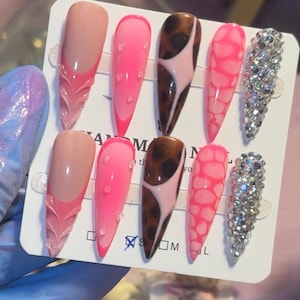 Op de afbeelding: Een set van acht lange, amandelvormige kunstnagels. Ontwerpen omvatten een French tip met een getextureerd accent, roze verlopen met hartaccenten, bruine en roze wervelingen, roze webpatronen en een volledig met strass bezette nagel.