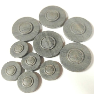 Può includere: Una collezione di bottoni circolari grigi con un design concentrico in rilievo. I bottoni, di varie dimensioni, sembrano essere fatti di un materiale solido, forse simile alla pietra. Sono disposti su una superficie bianca.
