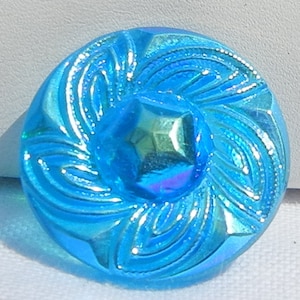 Könnte beinhalten: Ein runder, irisierender blauer Knopf mit einem zentralen, facettierten Edelstein. Der Knopf hat ein wirbelndes, geprägtes Design, das von der Mitte ausgeht. Die Farbe des Knopfes wechselt mit dem Licht und zeigt eine Reihe von Blautönen und violetten Farbtönen.