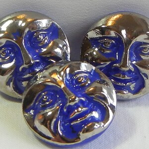 May include: Three silver-coloured buttons with a blue face design. The buttons have a shiny, metallic finish and a detailed depiction of a smiling face with blue accents. The buttons are likely for sewing or crafts.
