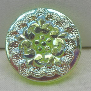 Puede incluir: Un botón redondo e iridiscente con un diseño floral detallado. El botón tiene un grupo central de pequeños círculos rodeados de hojas estilizadas y un borde festoneado. El color cambia entre verde, amarillo y toques de azul.