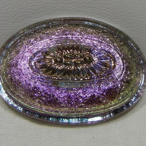 May include: Oval-shaped iridescent glass button with a floral design in the center. The button has a metallic sheen with colors ranging from purple to gold. The edges are slightly raised, and the button has a vintage aesthetic.