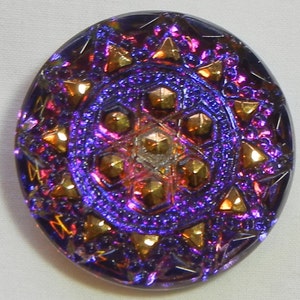 Könnte beinhalten: Ein runder, irisierender Knopf mit einem komplexen Design. Der Knopf hat ein zentrales Muster aus goldfarbenen geometrischen Formen und einen Rand aus Dreiecken. Die Farben wechseln von Lila zu Gold und Rot.