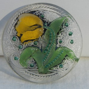 May include: A circular button with a silver finish, featuring a yellow tulip design with green leaves and stem. Small green embellishments are present. The button has an etched, decorative border.