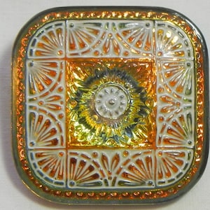 May include: Square, iridescent button with a detailed, Art Deco-inspired design. The button features a central floral motif surrounded by geometric patterns in shades of orange, yellow, and white, with a metallic gold border.