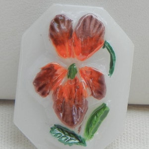 May include: A white, octagonal-shaped decorative piece featuring a raised, colorful flower design. The flower has orange and brown petals, a green center, and green leaves. The piece is likely made of resin or a similar material.