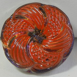 May include: A round, iridescent button with a spiral design. The button is primarily orange with hints of purple and blue. The button has a textured surface with a detailed pattern, and a raised edge.
