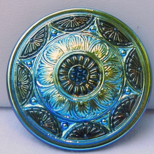 May include: A round, iridescent button with a detailed, embossed design. The button features a central floral pattern surrounded by ornate, fan-like shapes and a scalloped edge. The colors shift from blue to green.