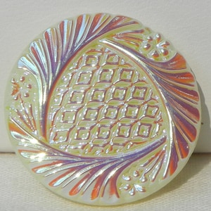 Peut inclure: Bouton rond irisé avec une surface texturée. Le bouton présente un motif central en losanges encadré d'un motif tourbillonnant avec des accents floraux. Les couleurs incluent le jaune, l'orange et le violet.